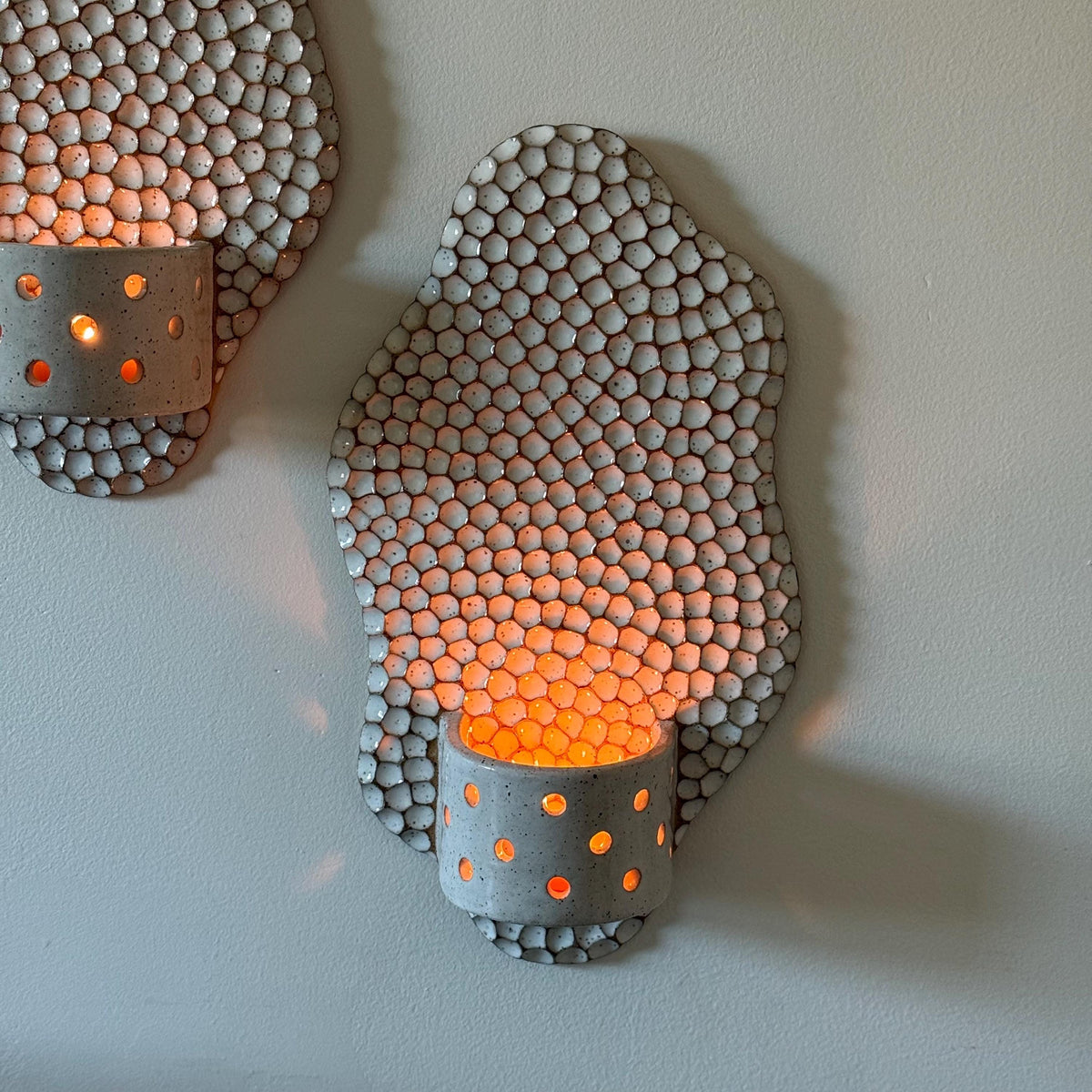 Wavy ceramic wall candle holder with white honeycomb texture and a perforated cup, glowing on a wall.