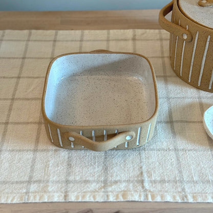 Square striped baking dish on checked linen beside the matching lidded pot.