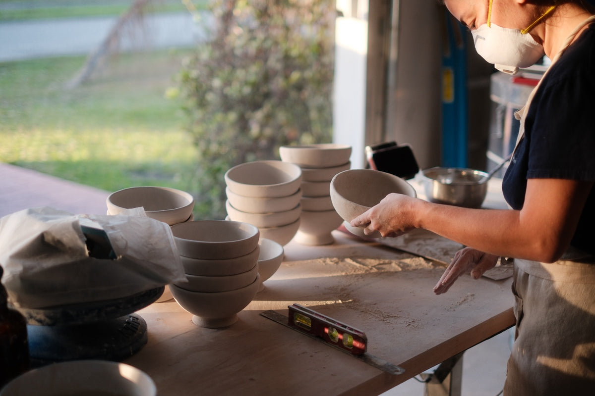 Mine finishing a handmade ceramic bowl beside stacks of bowls in her sunlit studio